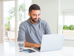 A man sits at his kitchen bench and smiles at his laptop