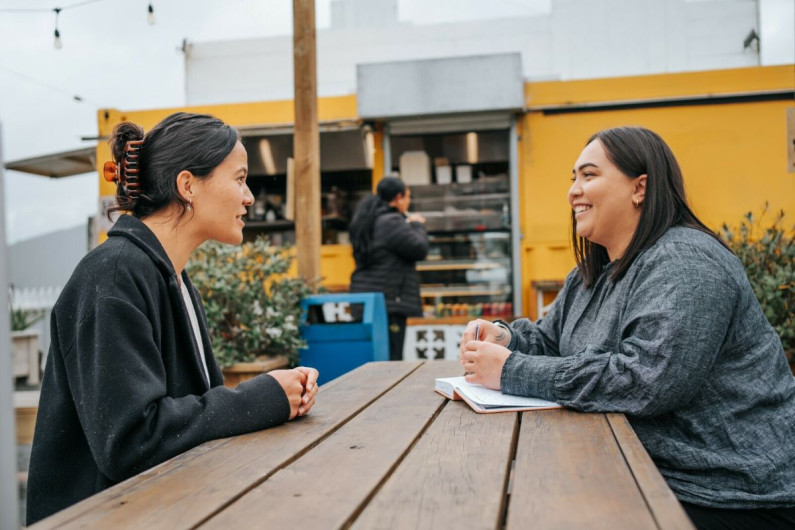 Two women talking at a picnic table outside in te reo Maori 