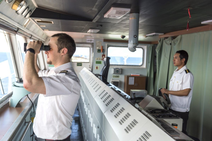 A ships master and a navigator on the bridge of a large ship, one is steering and the other is looking through binoculars 