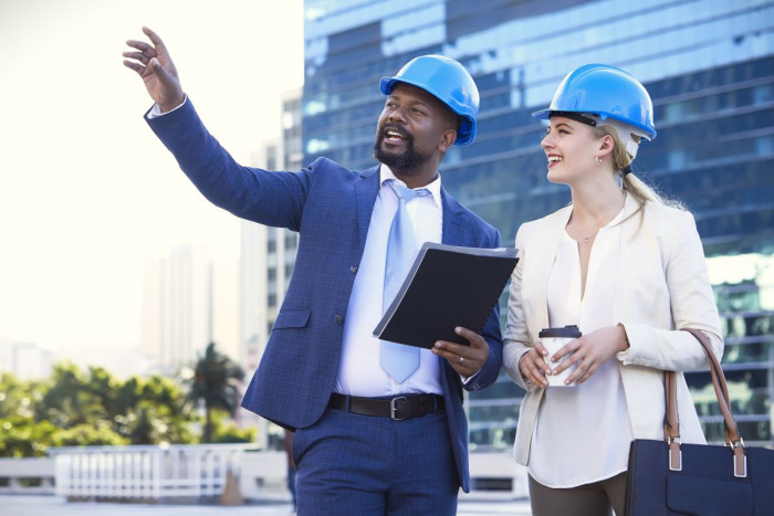 A man and a woman in business clothes and hard hats stand outside a multistorey building holding papers. They are both looking where he is pointing